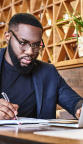 Portrait of dark-skinned businessman in dark-blue jacket and glasses sitting at the table, indoors, making notes, working with his laptop computer. Young African entrepreneur is coming up with new ideas. Modern interior