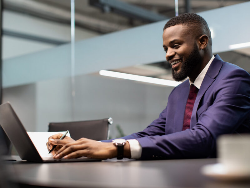 Cheerful african american young man in suit entrepreneur signing papers, working on laptop at office, side view, copy space. Happy black businessman taking notes while surfing on Internet
