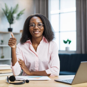 Smiling african american woman in eyeglasses and pink shirt sitting at desk with books and notes and showing thumbs up. Female tutor working at home during distance learning.